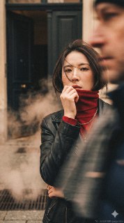 Prompt👇——超逼真的电影感肖像，高端时尚9：16画幅杂志风格摄影。发型：短发，发丝被风吹拂，飘散在脸上，以时尚的方式部分遮挡面部特征。主体：仅使用上传的面部参考图像作为主要拍摄对象。面部特征、骨骼结构和自然肤质（可见毛孔）需100%匹配。不得改变种族或性别特征。服装：黑色复古皮夹克搭配红色高领细线针织（堆叠领口），佩戴金色项链，优雅现代的造型。场景/环境：时间为冬天，街上有供暖热蒸汽，充满欧洲风格，一个复古公寓的门，门打开着，里面黑漆漆，动作/动态：前景有人快速移动，动态模糊（长曝光效果）挡住部分画面